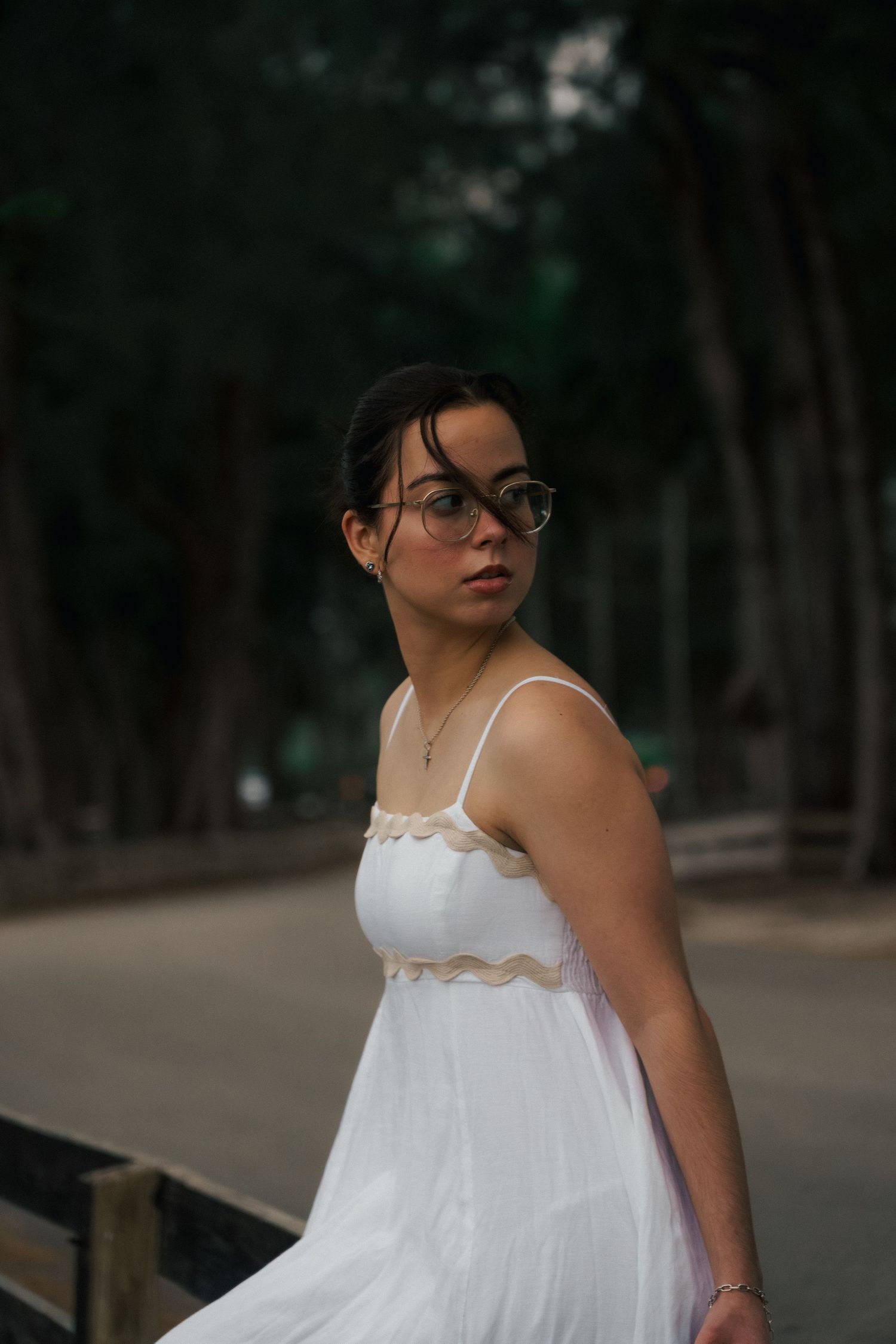 Young woman in white sundress and glasses poses outdoors near trees on a path.