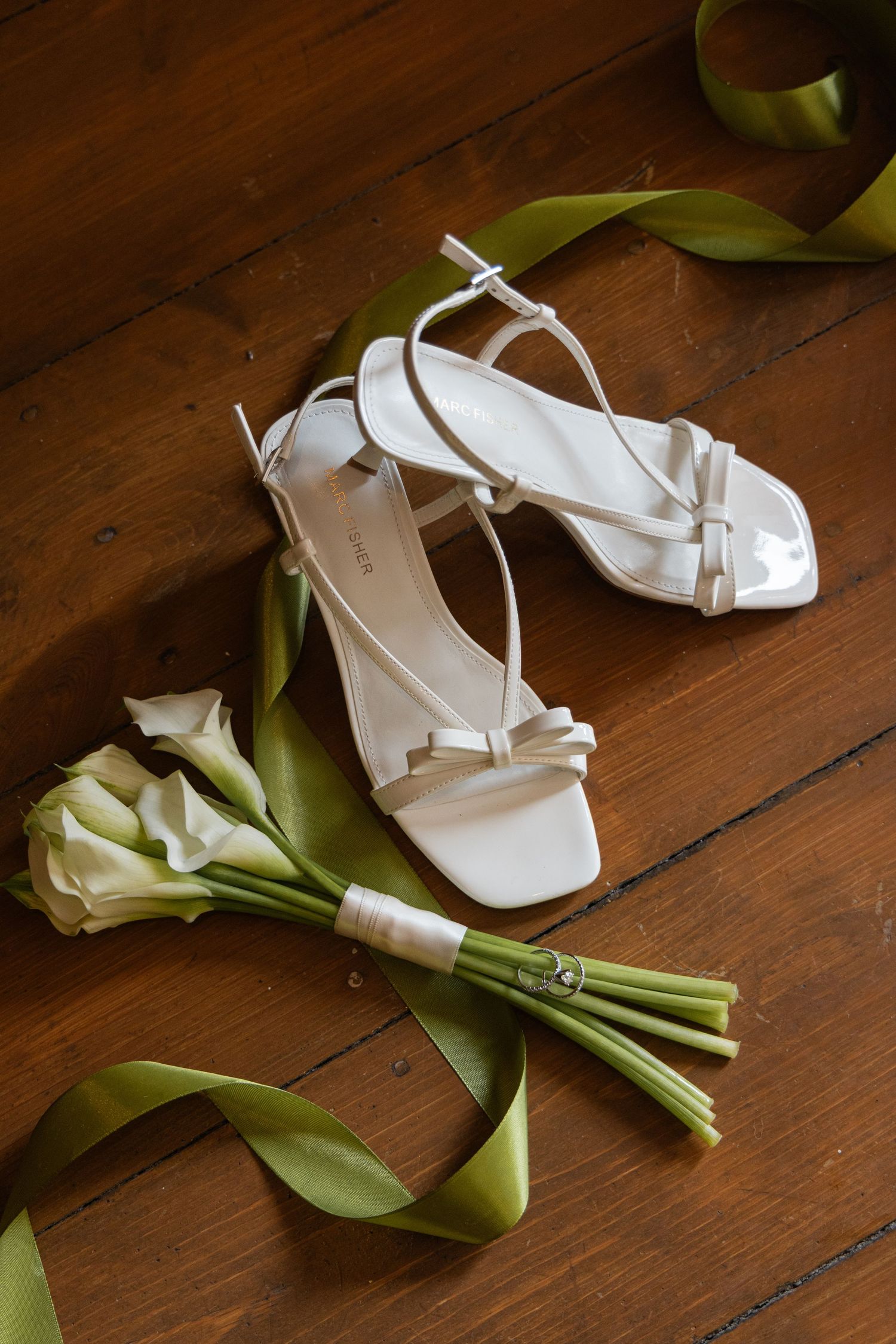 White strappy bridal heels beside white calla lily bouquet with green ribbon on rustic wooden floor.
