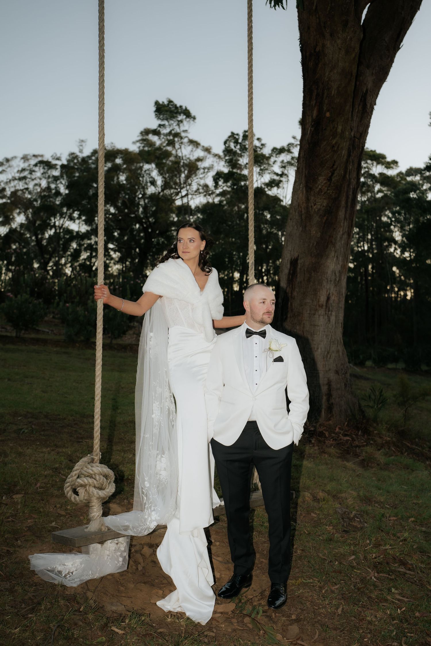 Bride standing on rope swing beside groom in white tuxedo jacket outdoors at dusk near large tree.