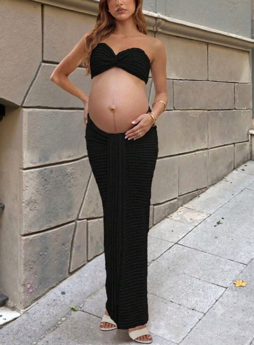 Pregnant woman in black strapless crop top and ribbed maxi skirt outfit, posing outdoors against stone wall.