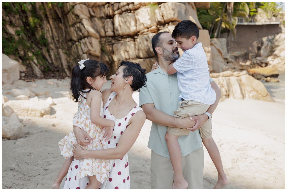 Family of four sharing a tender moment on a sandy beach, with parents holding young children near rocky cliffs.
