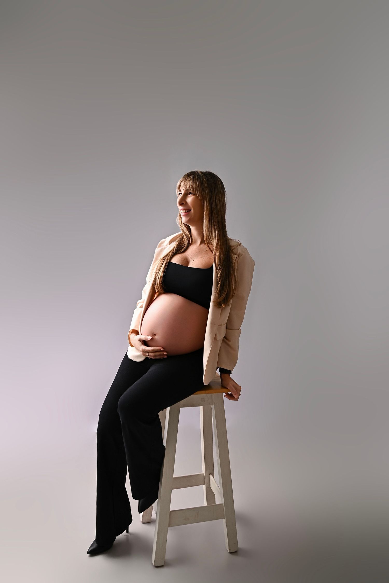Pregnant woman in black crop top and trousers with beige blazer sitting on stool, cradling baby bump in studio.
