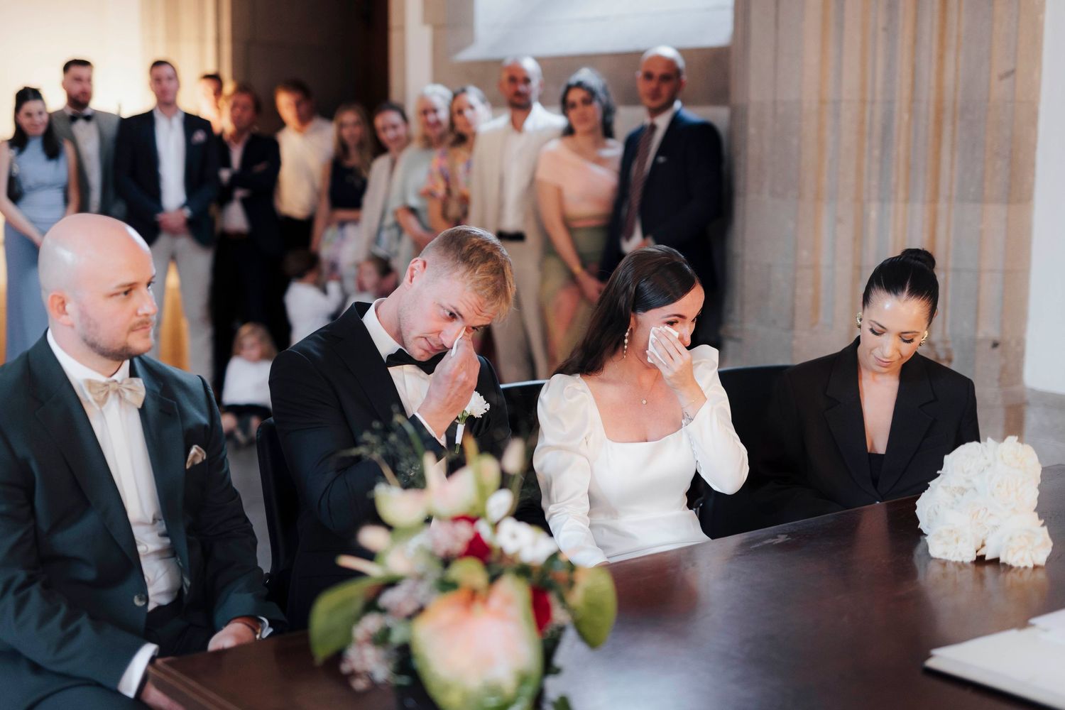 Emotional bride and groom wiping tears during wedding ceremony at elegant venue with guests in background.