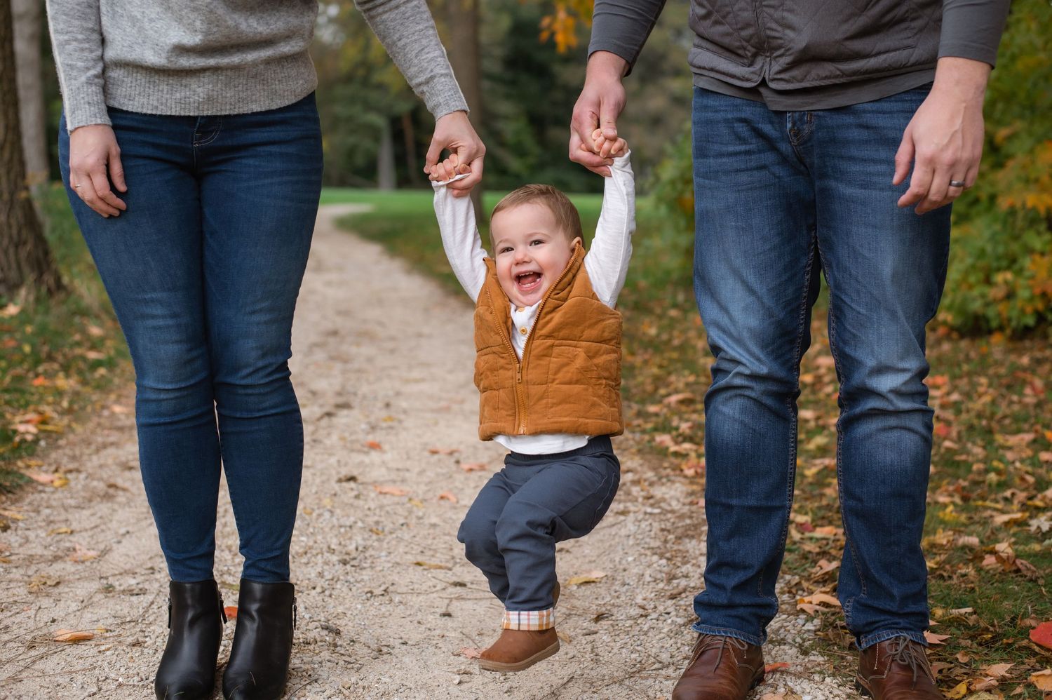 Happy baby taking first steps on a tree-lined path, held by parents hands outdoors in autumn.