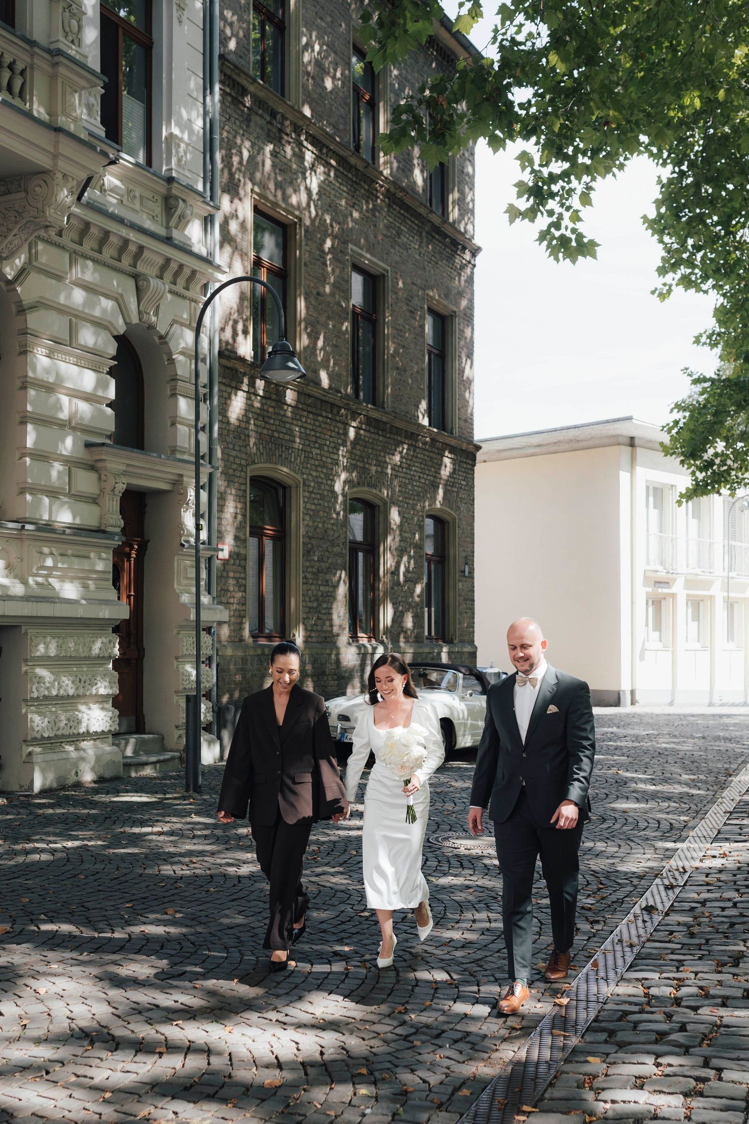 Eine Braut im weißen Kleid läuft an ihrem Hochzeitstag mit den Trauzeugen im historischen Köln zu ihrem First Look. Oldtimer im Hintergrund.