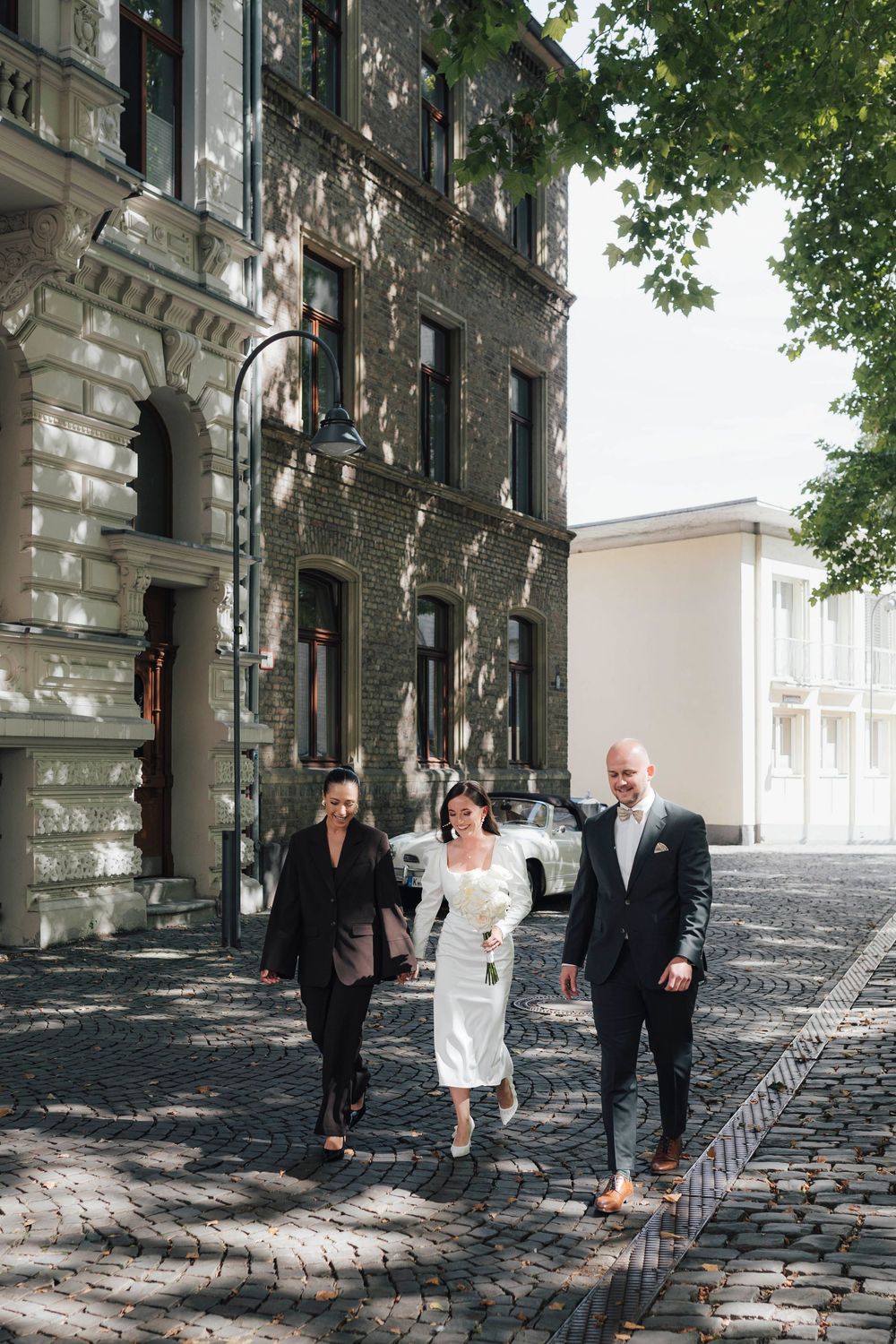 Bride in white dress walks with two guests on cobblestone street past historic brick buildings on wedding day.