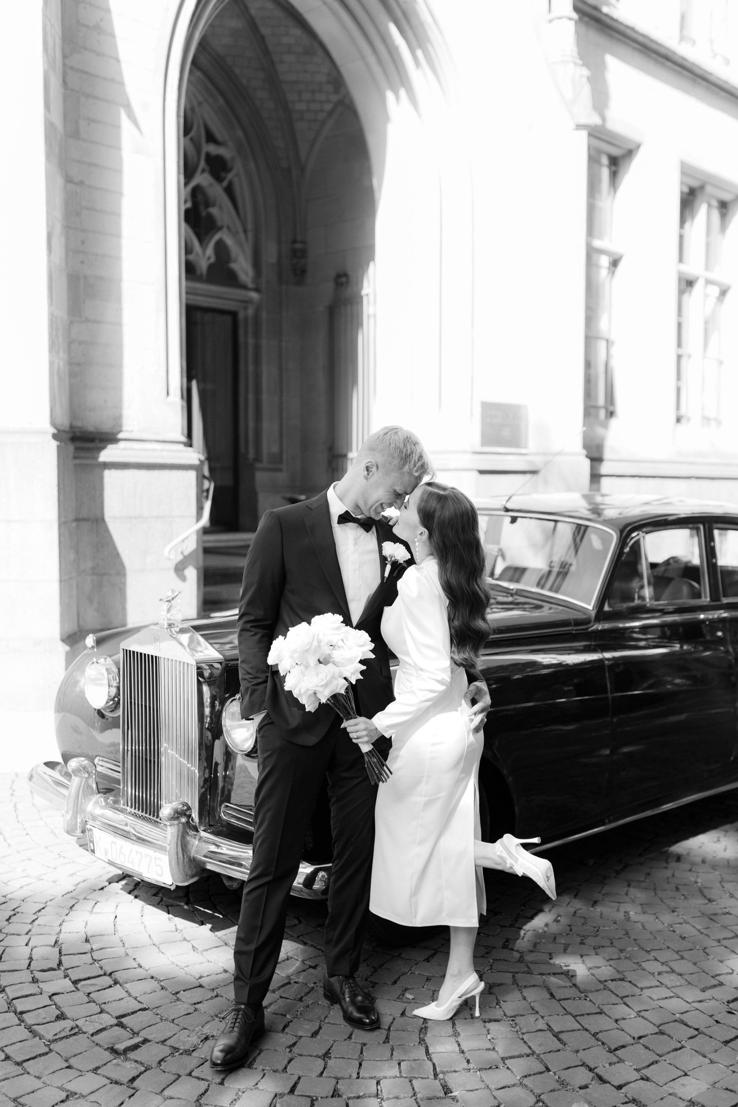 Black and white photo of elegant wedding couple kissing beside vintage Rolls-Royce, bride in white dress holding bouquet.