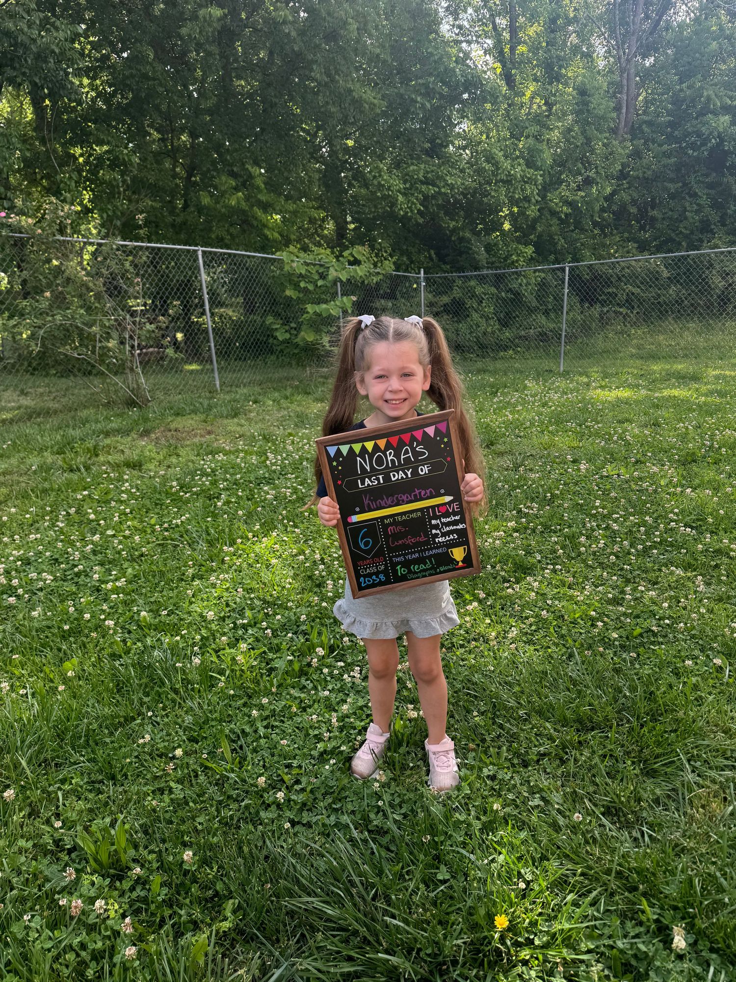 Young girl holding a chalkboard sign on last day of school, standing in green yard with chain-link fence.