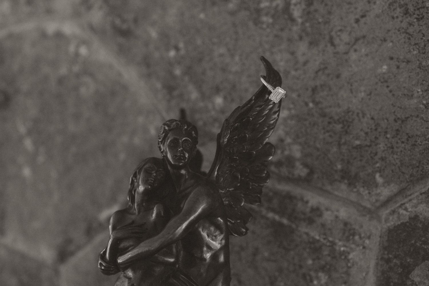 Black and white photo of a dark bronze angel statue with wings, holding a figure, against a stone wall.