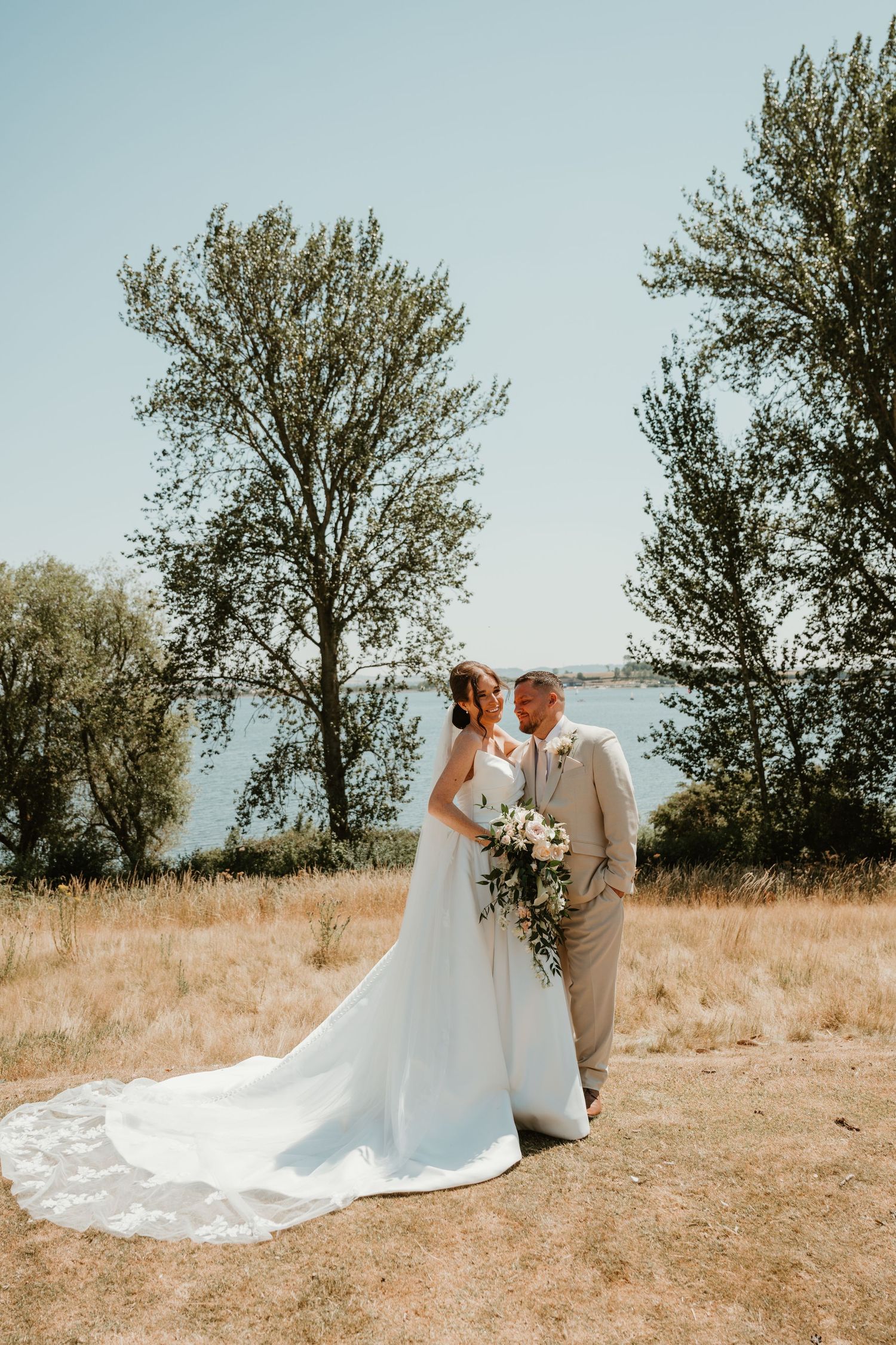 Bride and groom pose outdoors near a lake with trees and blue sky on their wedding day.