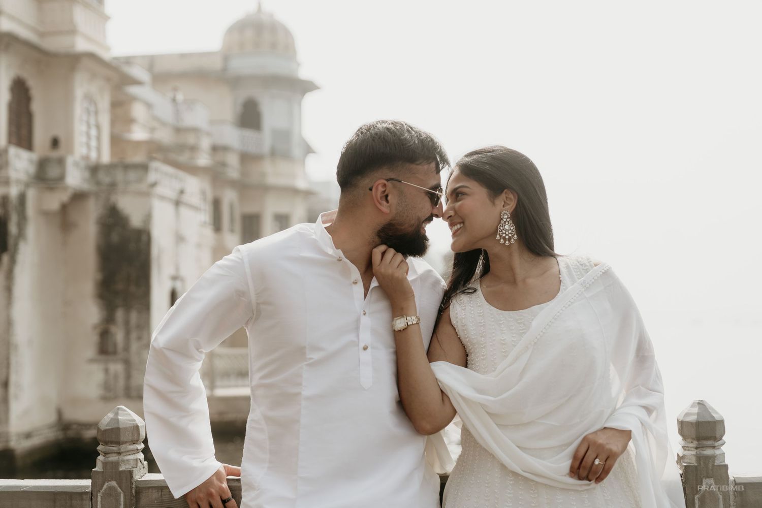 Couple in white traditional Indian attire sharing a romantic moment near a historic palace.