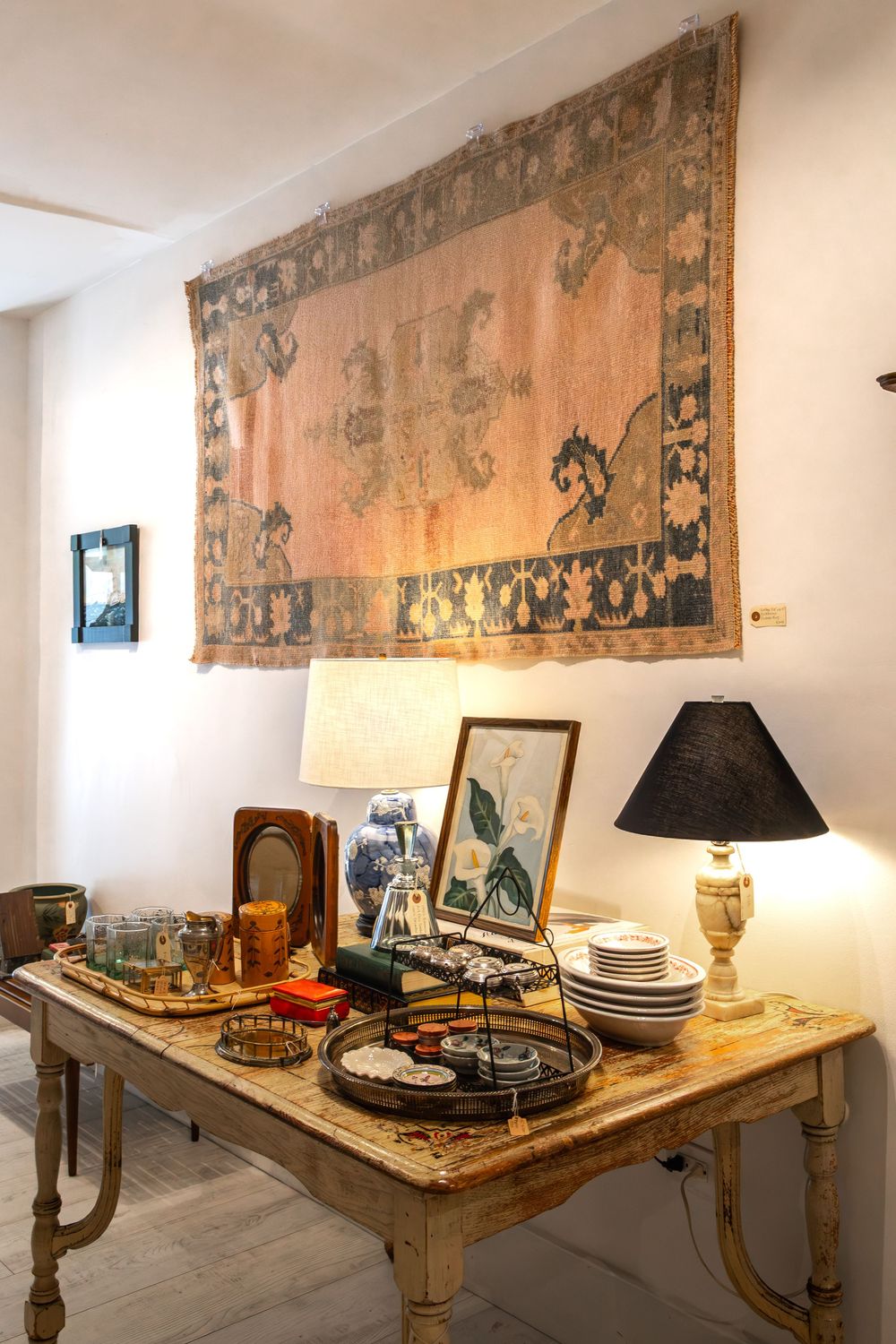 Antique wooden table displaying lamps, decorative items, and plates beneath a vintage tapestry on a white wall. Swell House La Grange