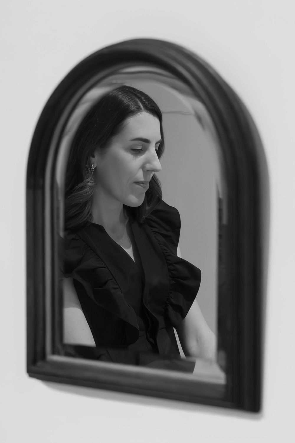 Black and white reflection of a woman in dark ruffled top gazing down, captured in an arched wall mirror. Swell House La Grange