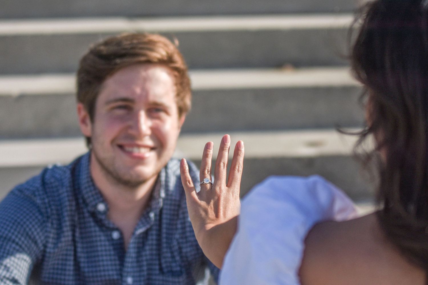 Smiling man on one knee showing engagement ring to woman during outdoor proposal.