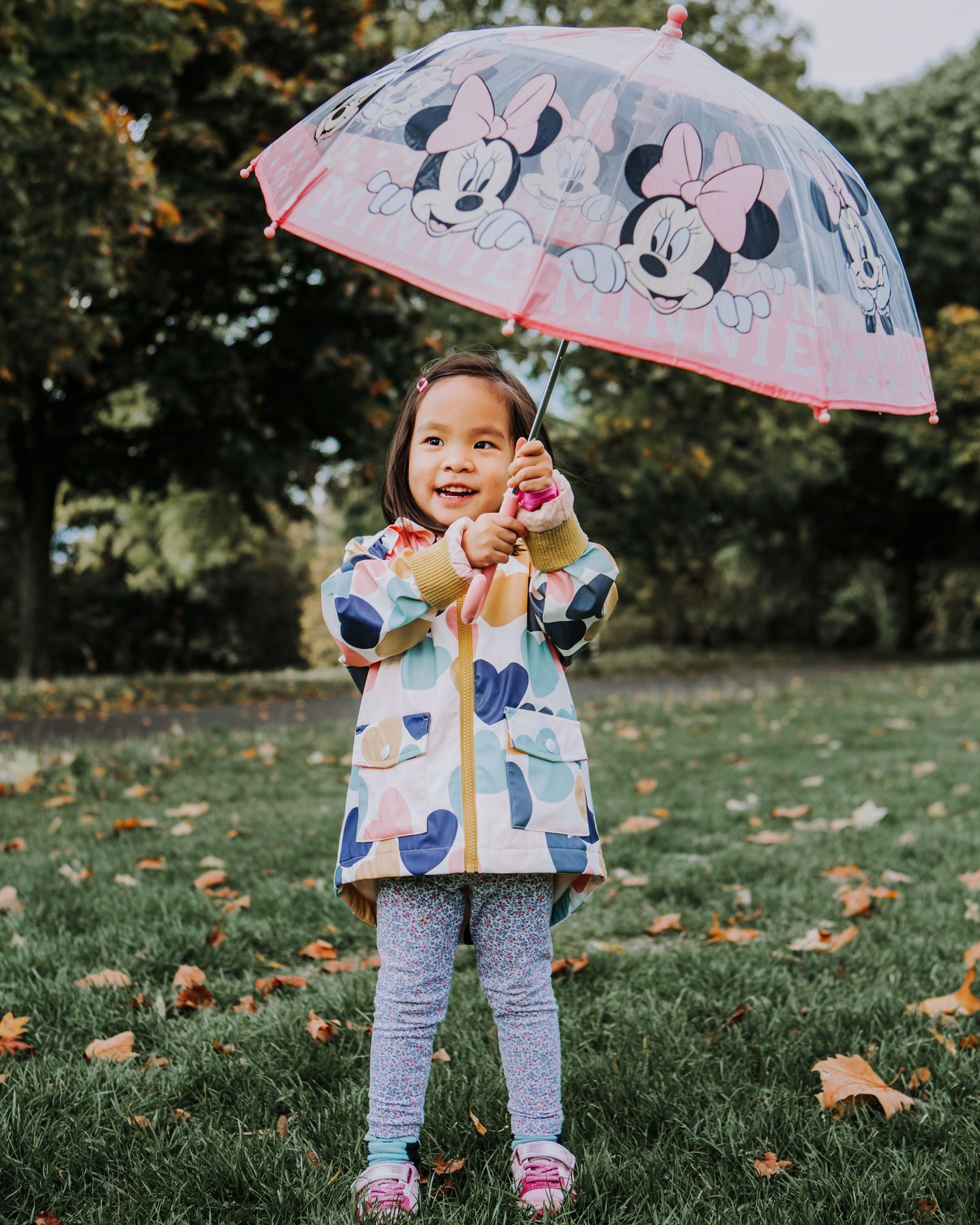 Little girl smiling while holding a colourful Minnie Mouse umbrella during an outdoor children's photography session.