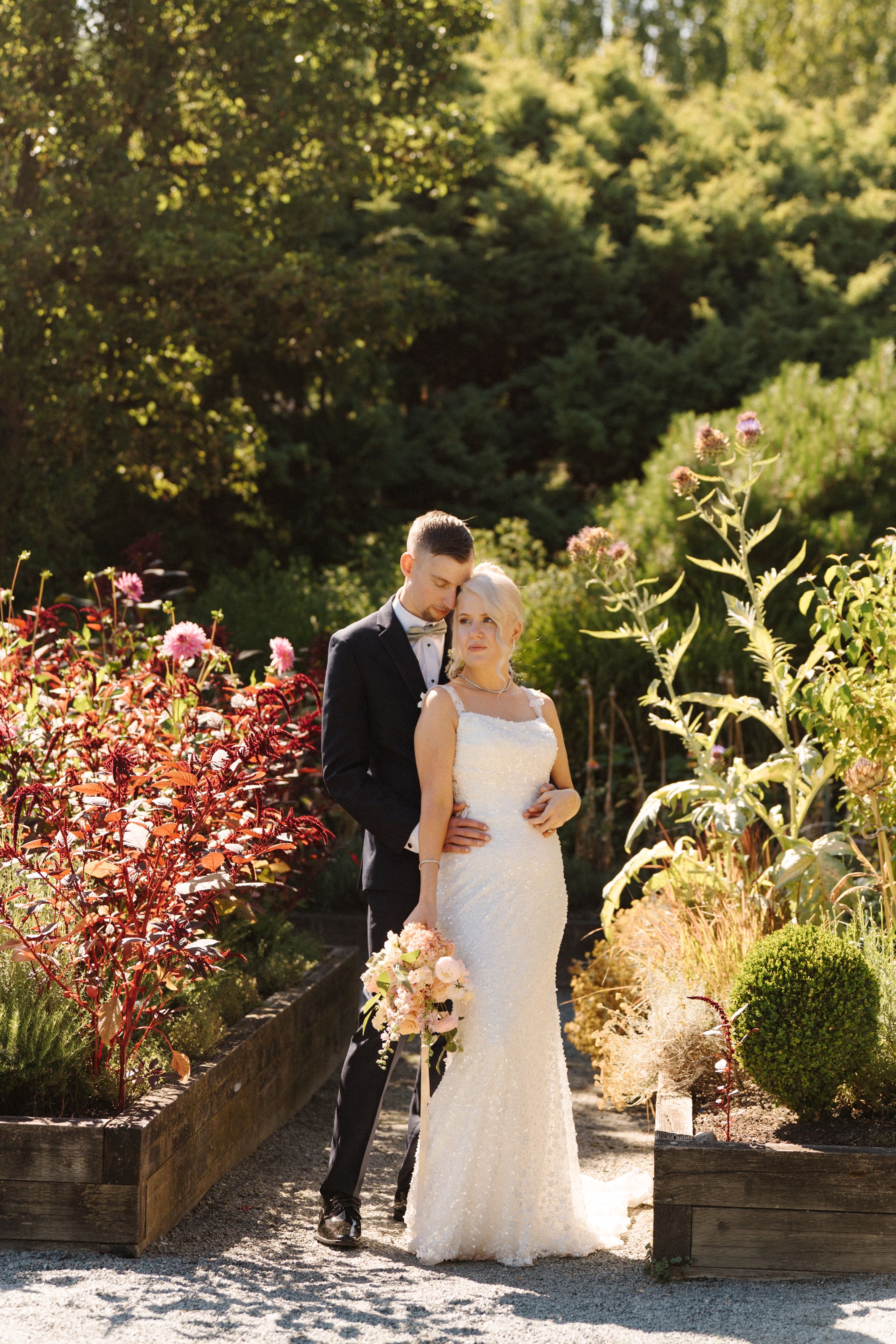Elegant bride and groom posing among colorful garden beds, bride holding a bouquet on their wedding day.