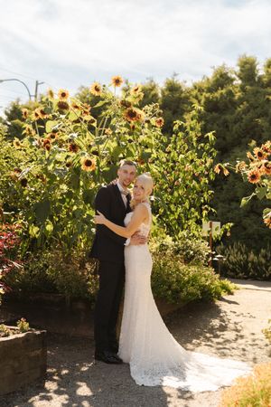 Bride and groom embracing in a sunlit garden with sunflowers blooming behind them on their wedding day.