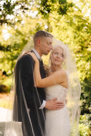Bride and groom embrace under a flowing veil in a sunlit garden setting.