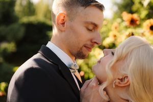 A groom in a dark suit and bow tie shares a romantic moment with a blonde bride outdoors.