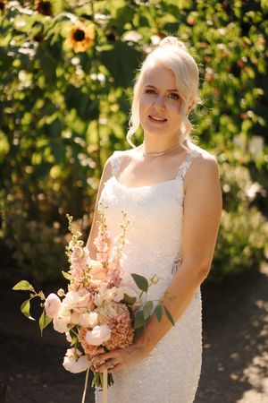 Blonde bride in white sequin gown holding pink floral bouquet with sunflowers in background.