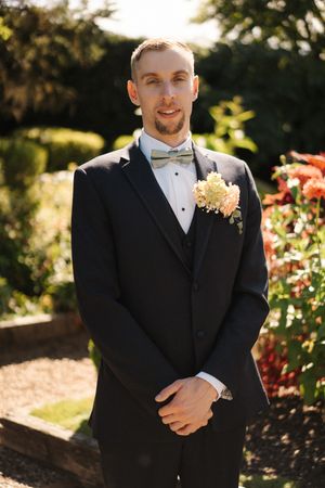 Groom in black tuxedo with bow tie and floral boutonniere posing in garden setting.