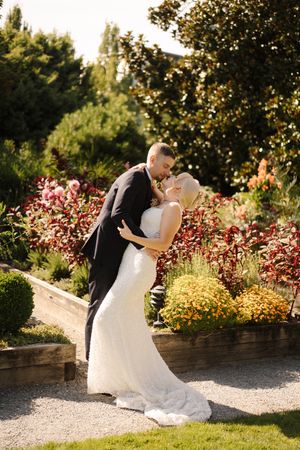 Bride and groom share a romantic kiss in a sunlit garden with colorful autumn flowers during their wedding day.