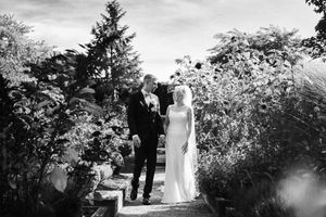 Black and white photo of bride and groom walking through lush garden on their wedding day.