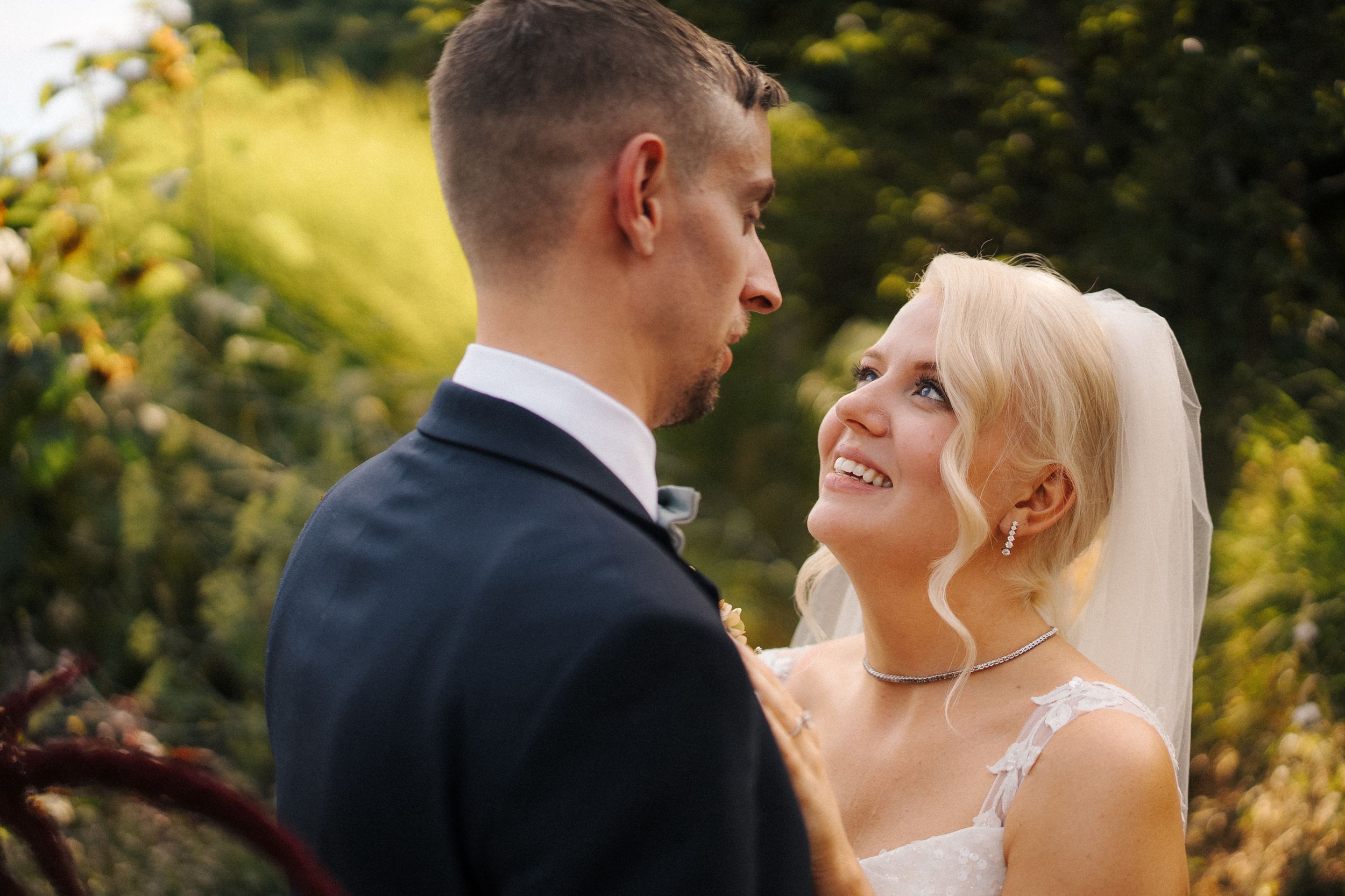 Smiling blonde bride in veil gazes at groom outdoors in golden sunlight.
