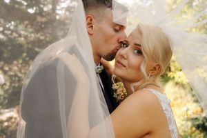 A bride and groom share a romantic moment under a flowing white veil outdoors on their wedding day.