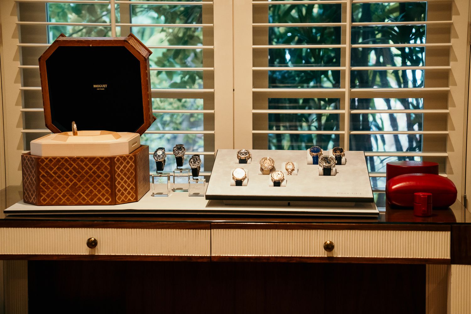 Elegant watch display on wooden desk with jewelry box and shuttered window backdrop.
