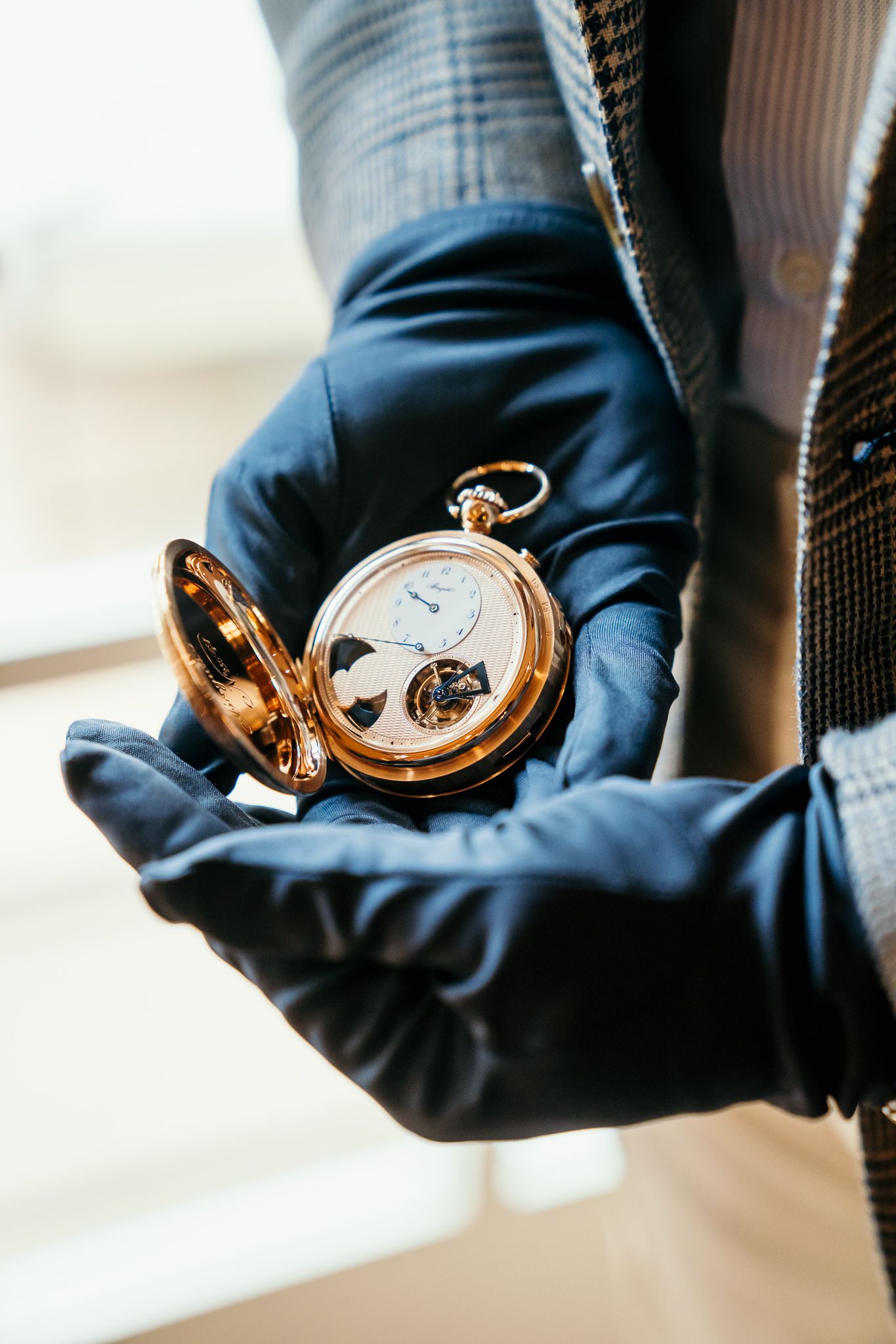Gloved hands holding open gold Breguet pocket watch with tourbillon movement.