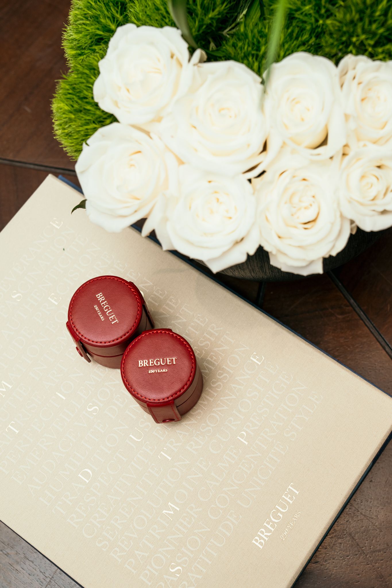 Two red Breguet watch cases on a branded book beside white roses arrangement.