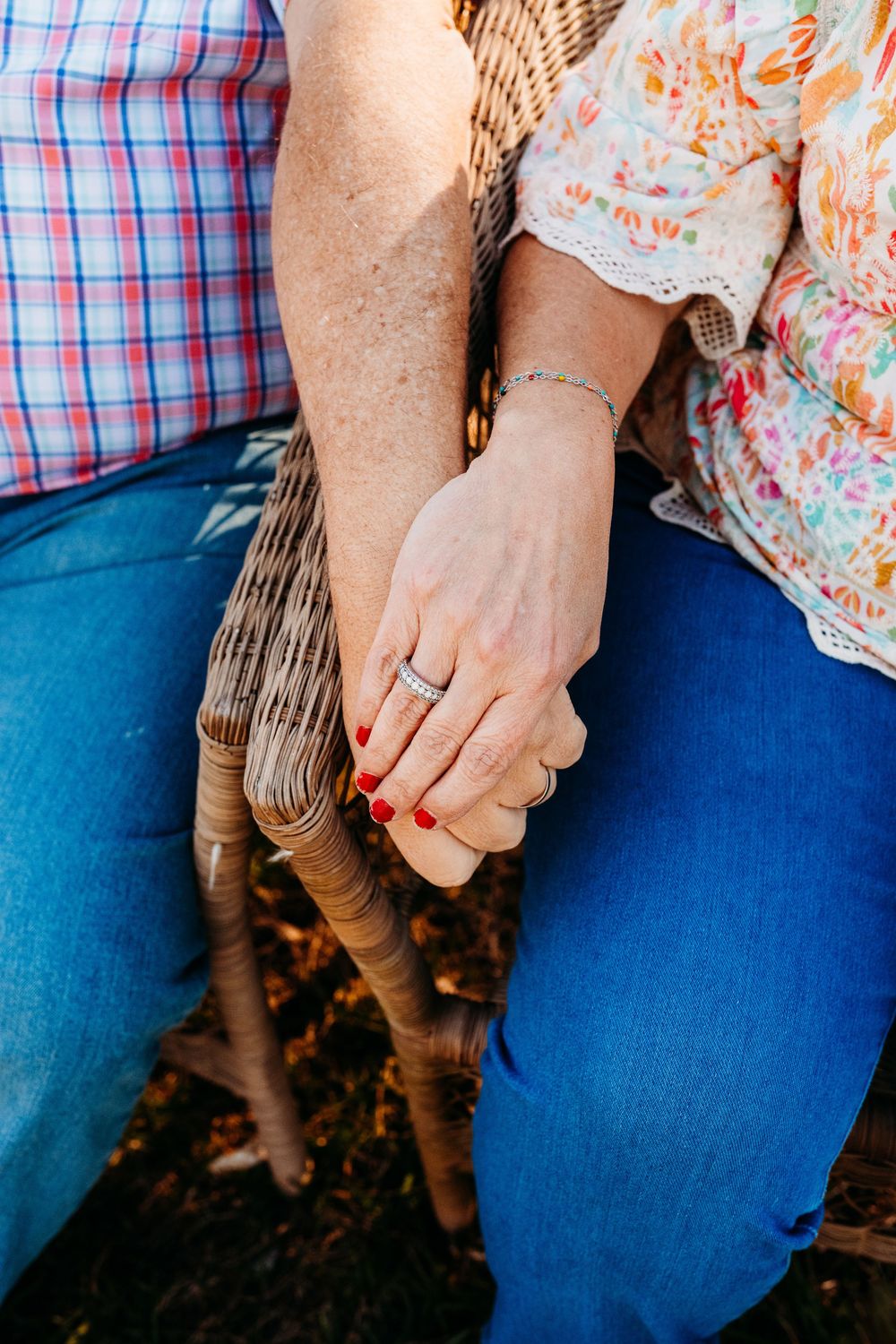 Close-up of an elderly couple holding hands, woman wearing a diamond ring and red nail polish, seated on a wicker chair.