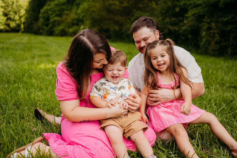 Happy family of four sitting on green grass, parents smiling at laughing toddler boy and girl in pink dress.
