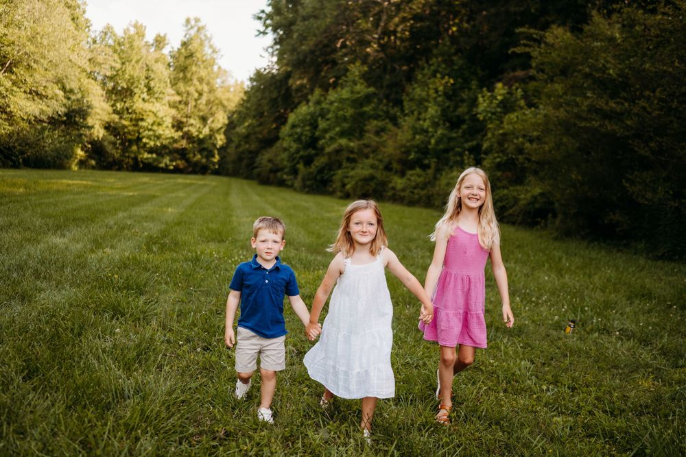 Three happy children holding hands and running through a lush green field during golden hour.