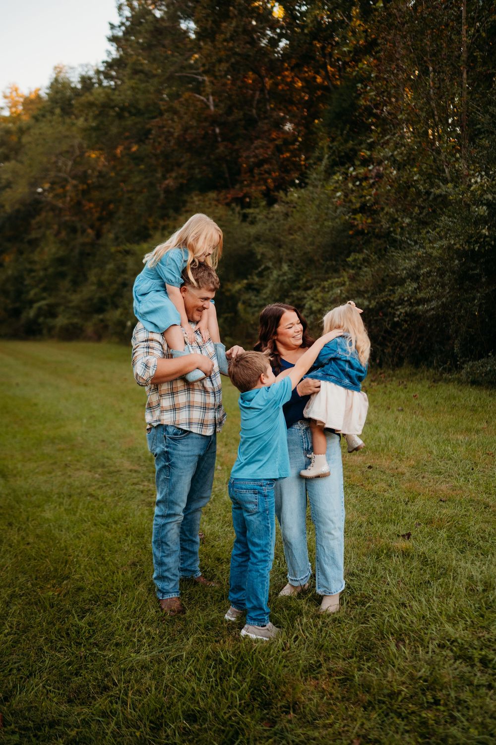 A happy family of five enjoys a playful outdoor moment in a lush green field, with children on shoulders and in arms.