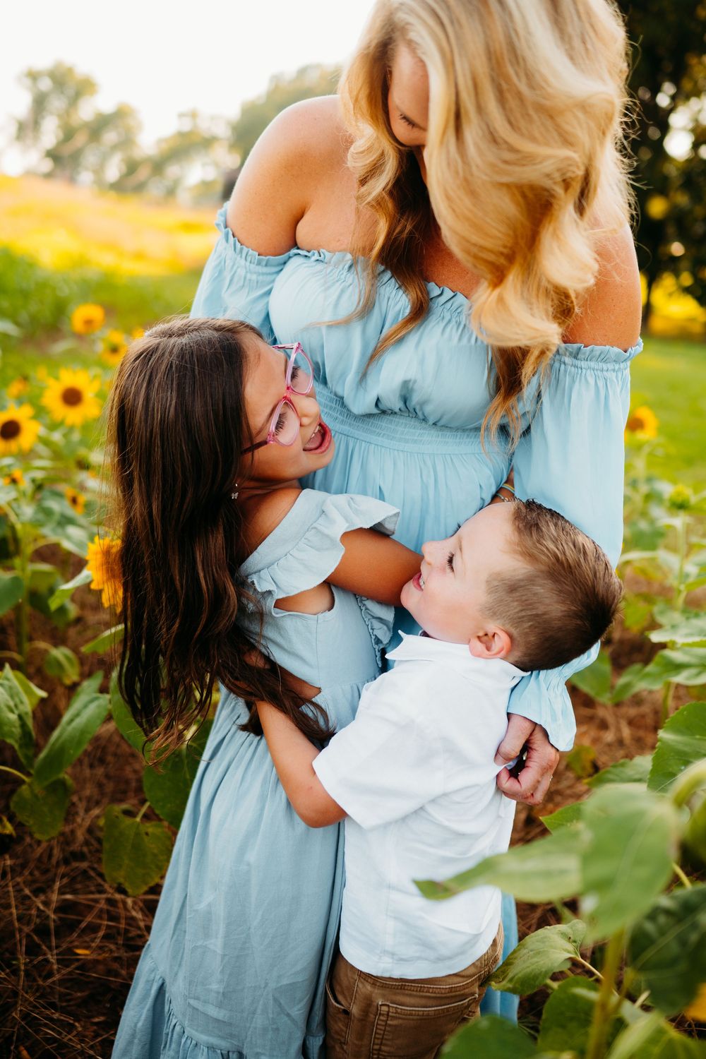 A mother in a blue dress hugs her daughter and son in a sunny sunflower field.