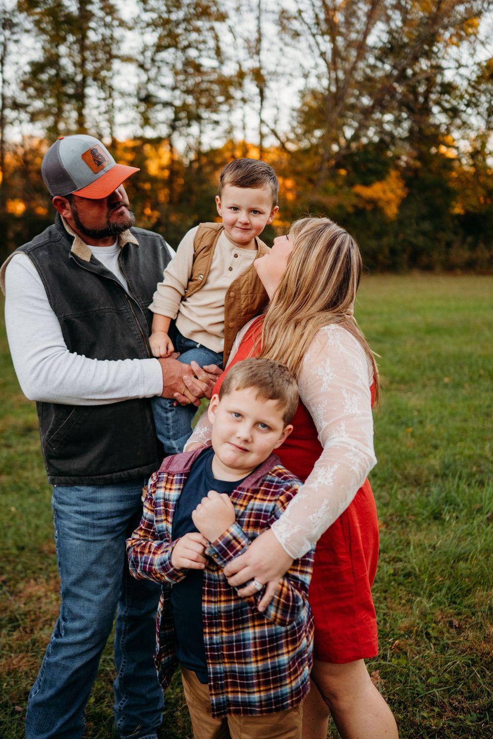 Family of four posing outdoors at golden hour, parents embracing two young boys in a green field with autumn trees.