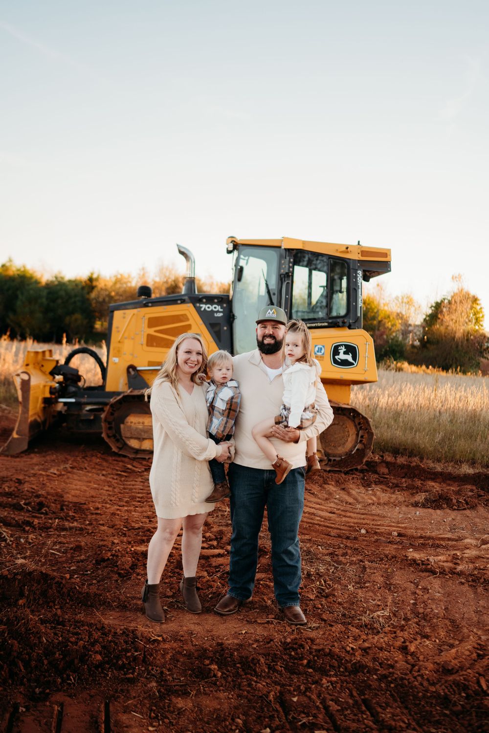 Family of four posing in front of a John Deere bulldozer on a red dirt construction site at golden hour.