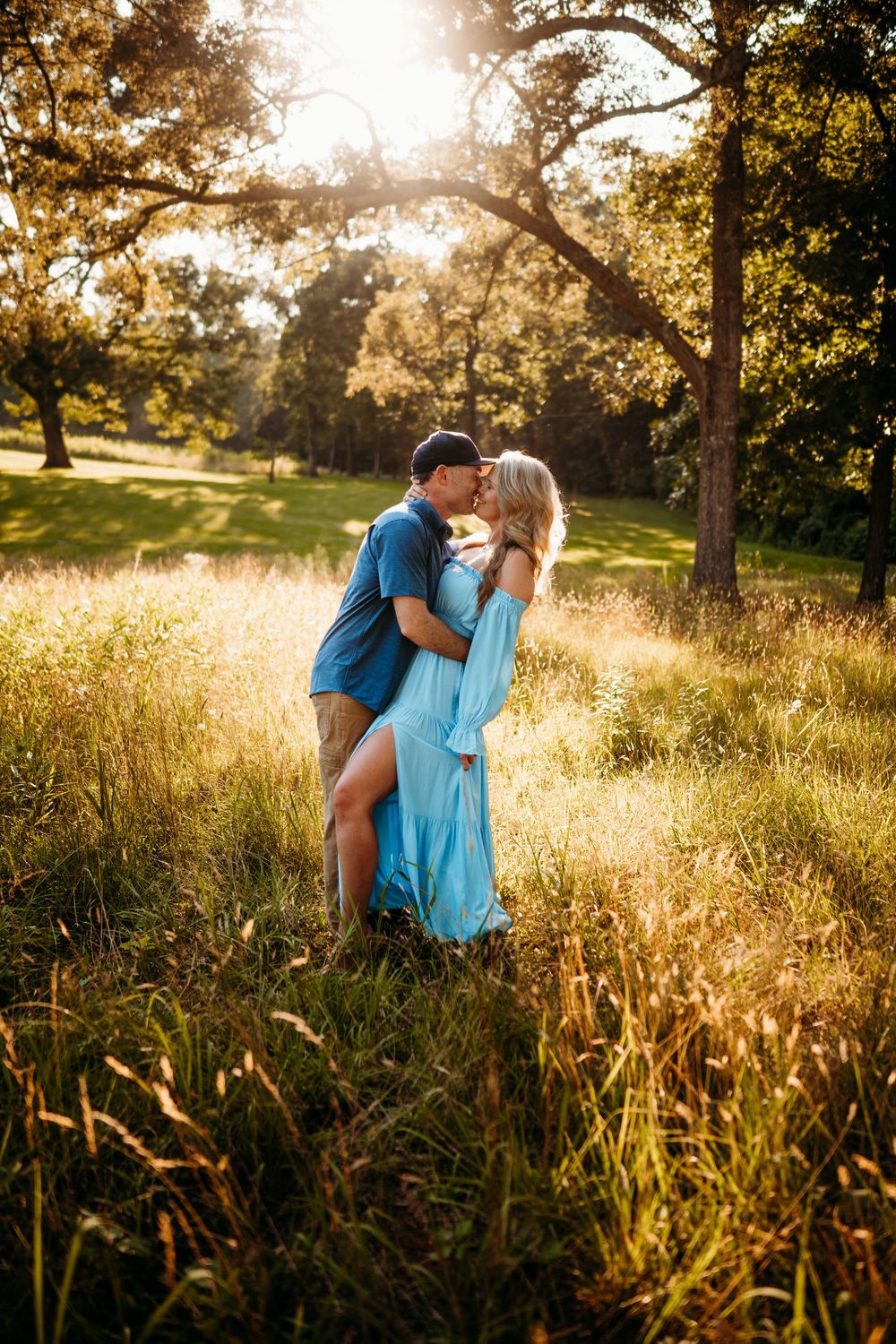 Couple sharing a tender kiss in golden sunlit meadow, woman in flowing blue dress surrounded by tall grass and trees.
