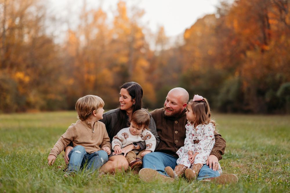 Family of five sitting on grass in autumn park, parents and three young children smiling together outdoors.
