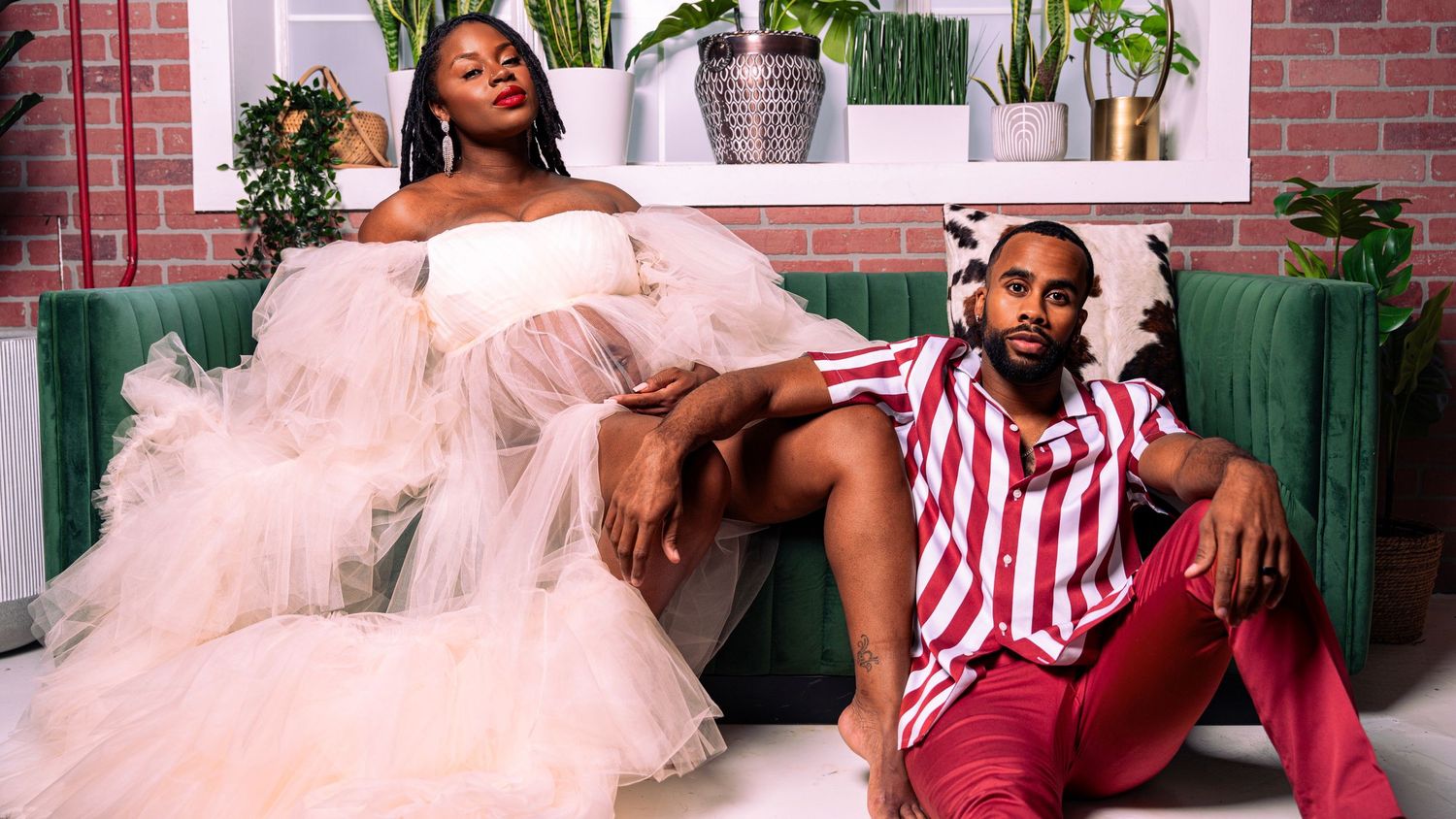 A woman in a white tulle gown and a man in a red striped suit pose together on a green velvet couch.