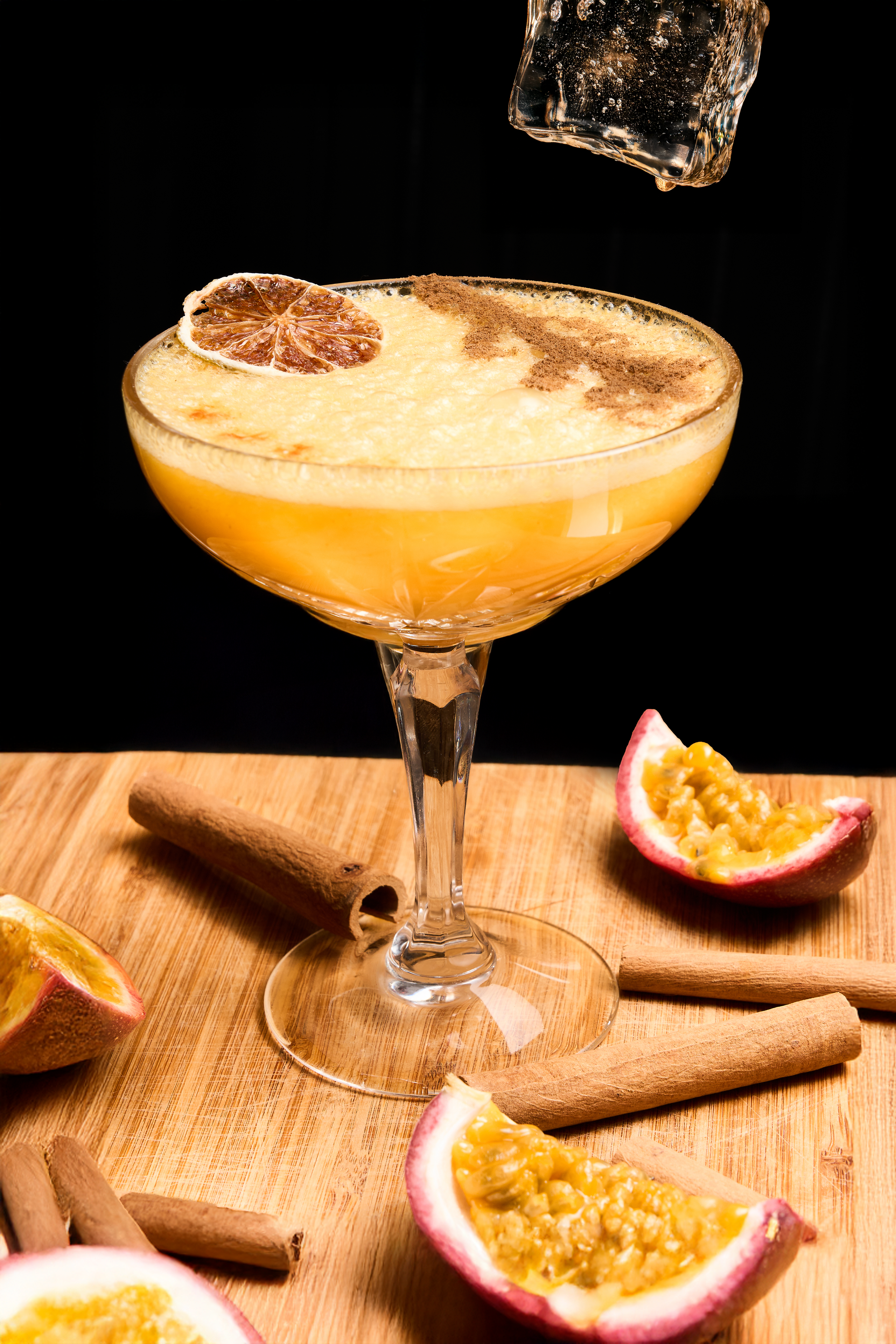 Golden passion fruit cocktail in coupe glass with cinnamon sticks, fresh passion fruit, and dried citrus garnish on wood board.