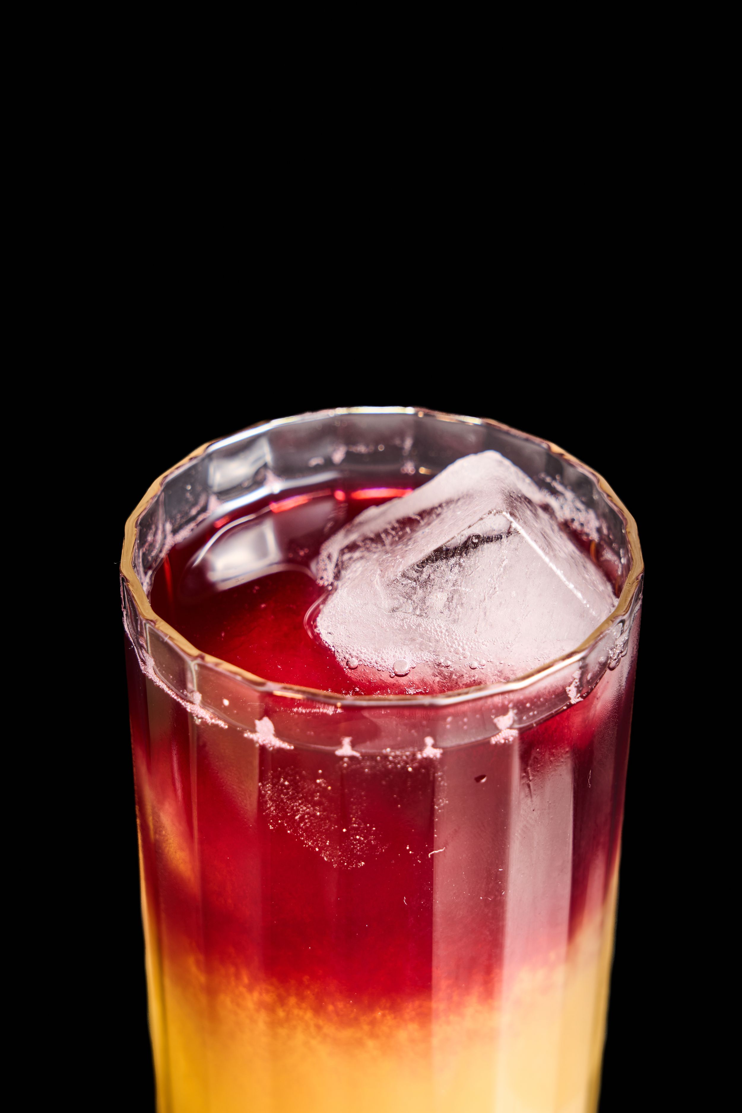 A layered cocktail with red and orange hues, topped with ice cubes in a tall glass on a black background.