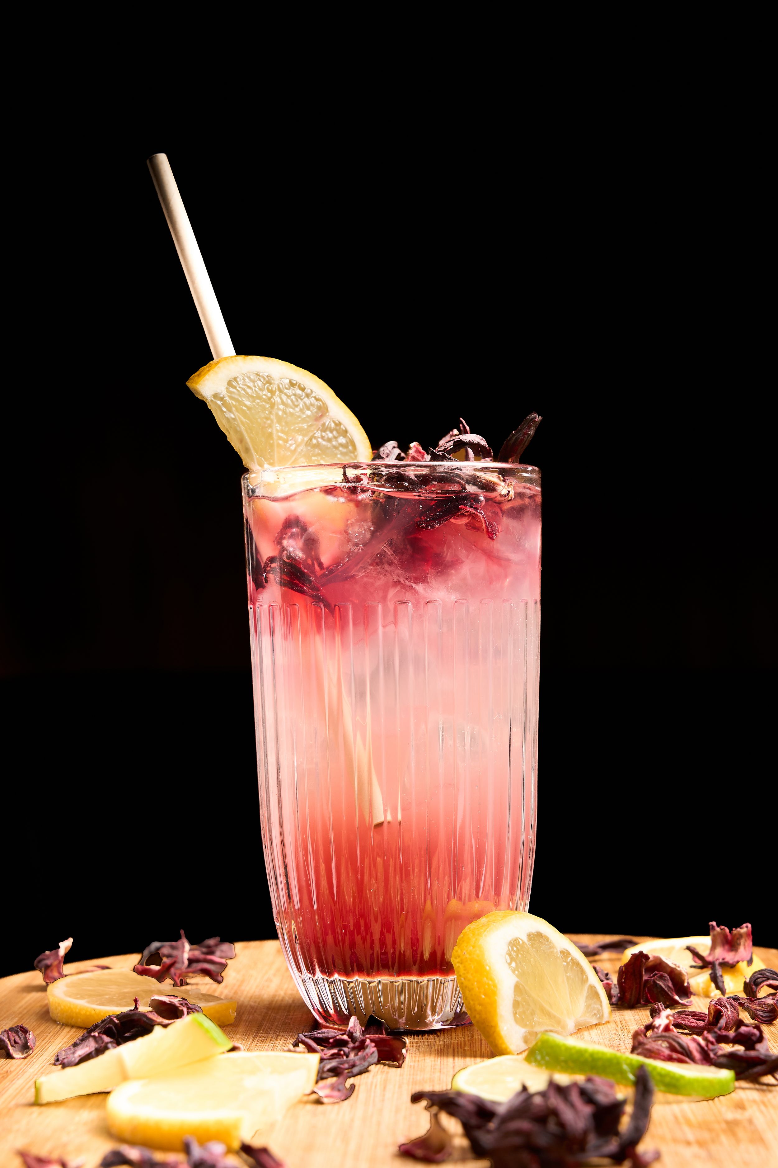 Pink hibiscus lemonade cocktail in ribbed glass with straw, lemon slice garnish, and dried flowers on wooden board.