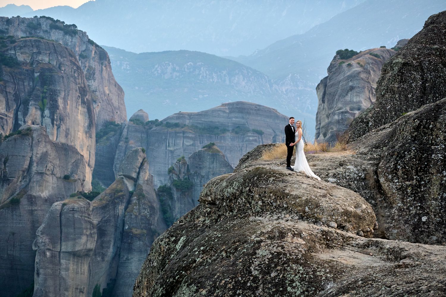 Wedding couple embracing on a rocky cliff in Meteora, Greece, with mountains in background.