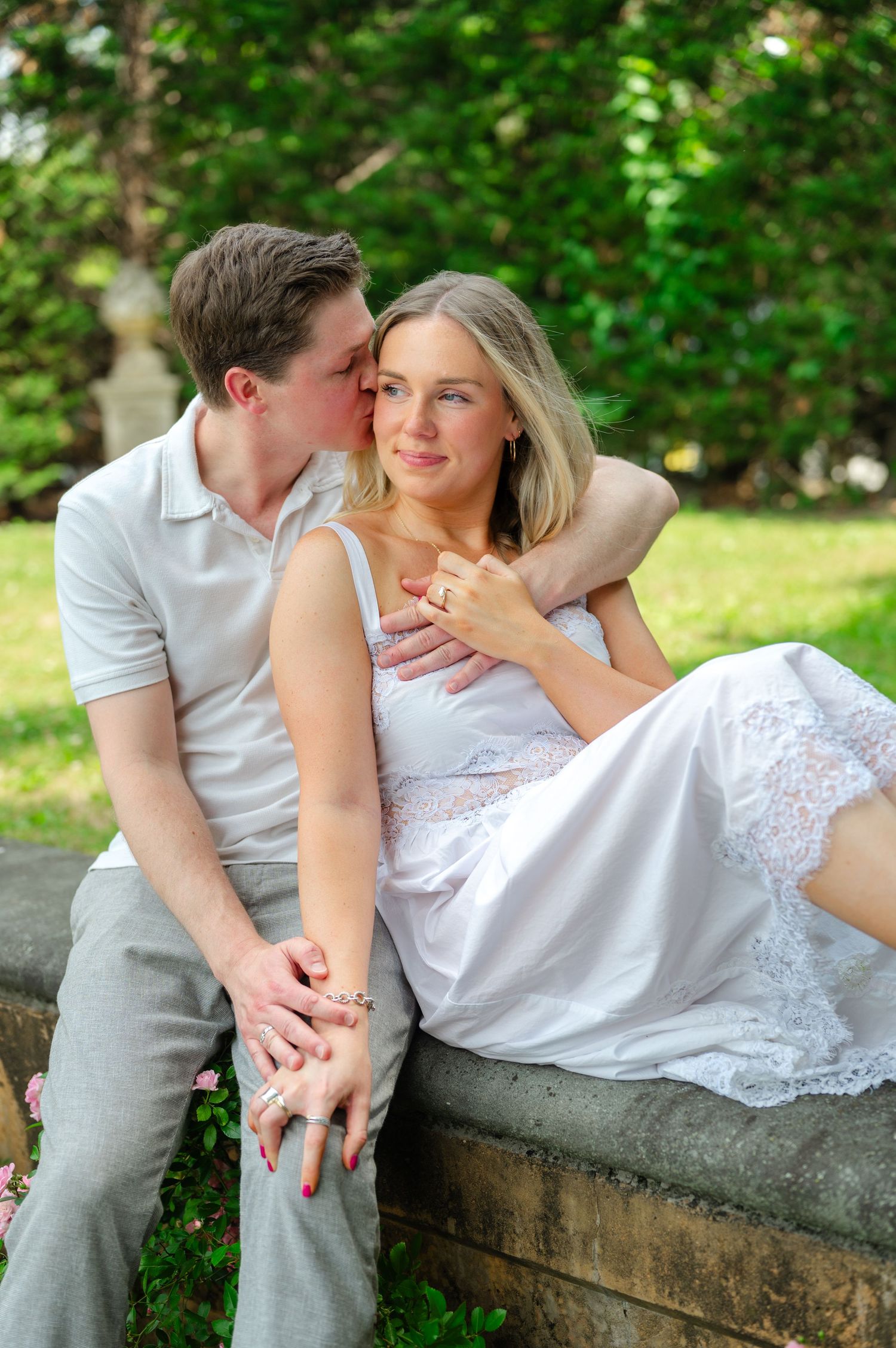 Couple sitting on stone wall outdoors, man kissing woman's cheek, she wears white lace dress, green trees behind them.