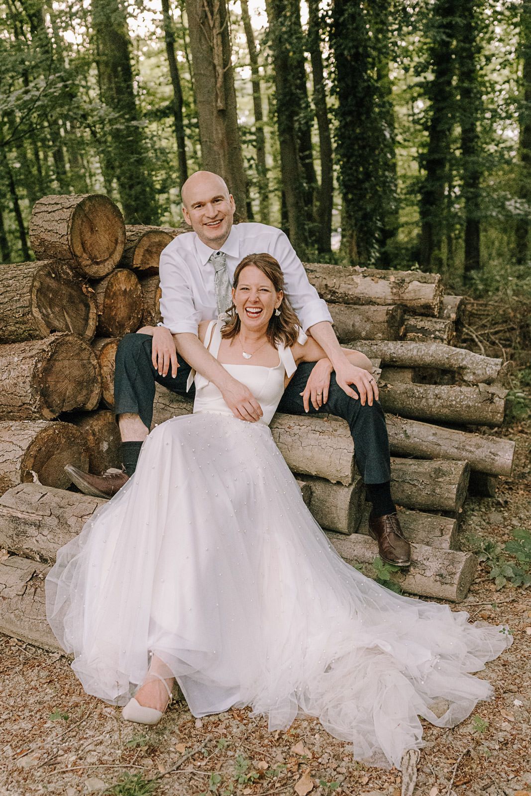 A smiling bride in a white gown and groom in a white shirt sit together on stacked logs in a lush forest setting.