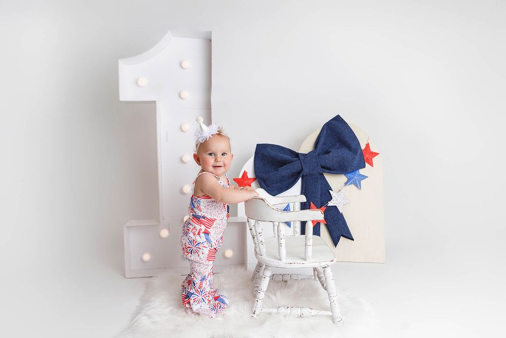 Baby girl in floral outfit stands by large number 1 prop with white rocking chair by affordable baby photographer in Riverside county.