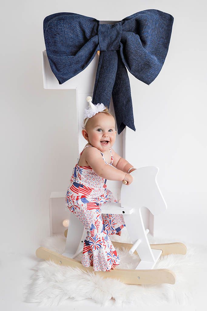 Baby girl in patriotic romper standing on white rocking horse by affordable baby photographer in Riverside county.